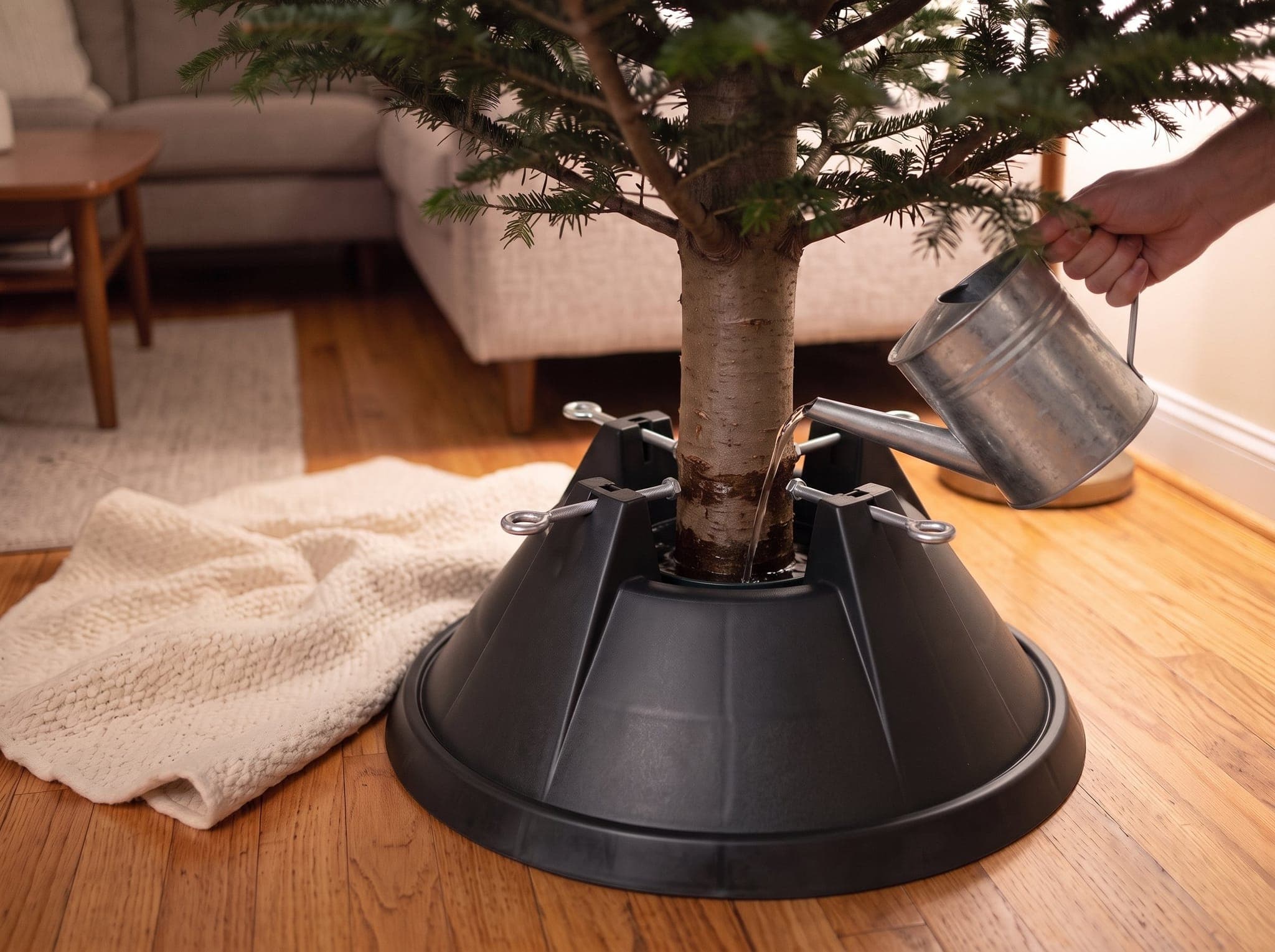 Water poured from a metal watering can into the reservoir of a molded plastic Christmas tree stand, four eye bolts gripping the fir trunk