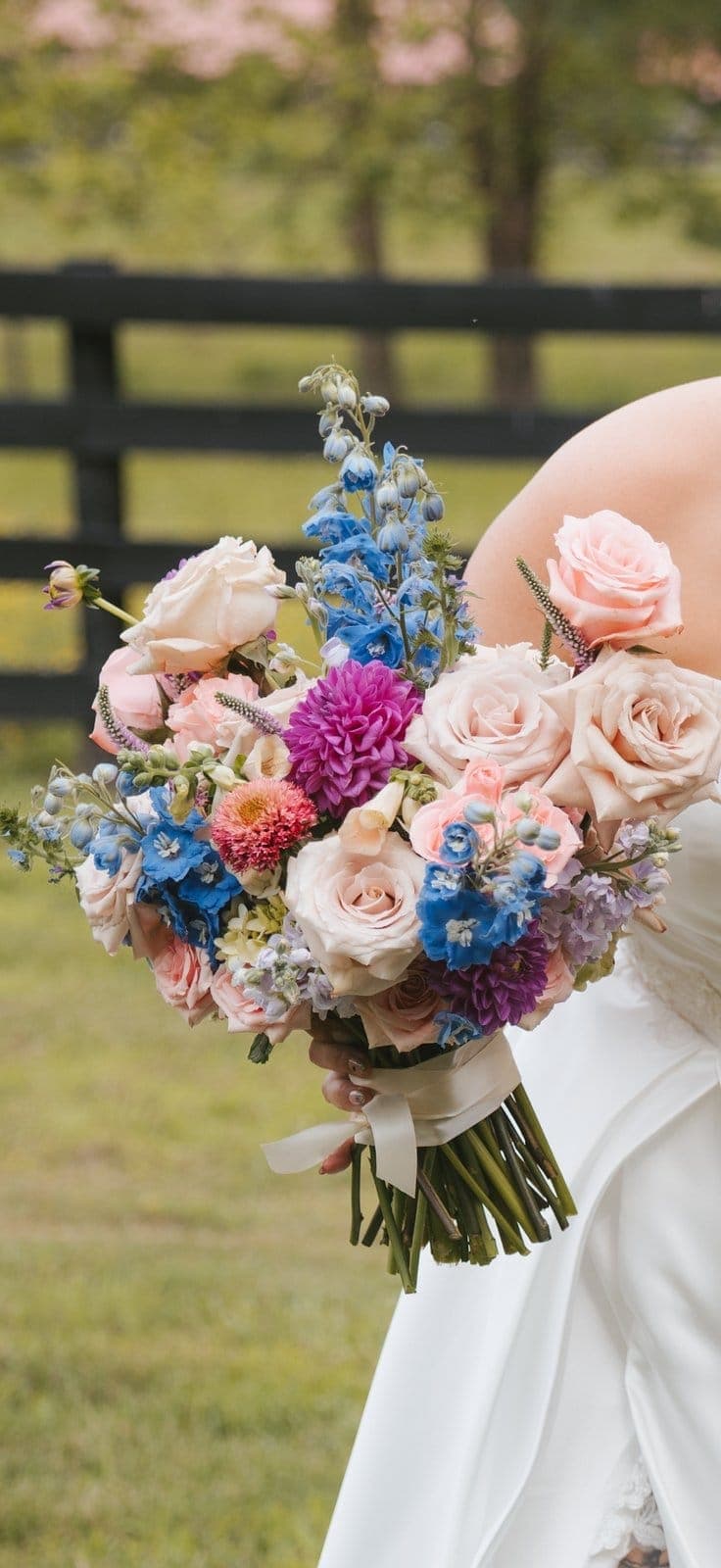 Bridal bouquet of blush roses, blue delphinium, and magenta dahlias held outdoors