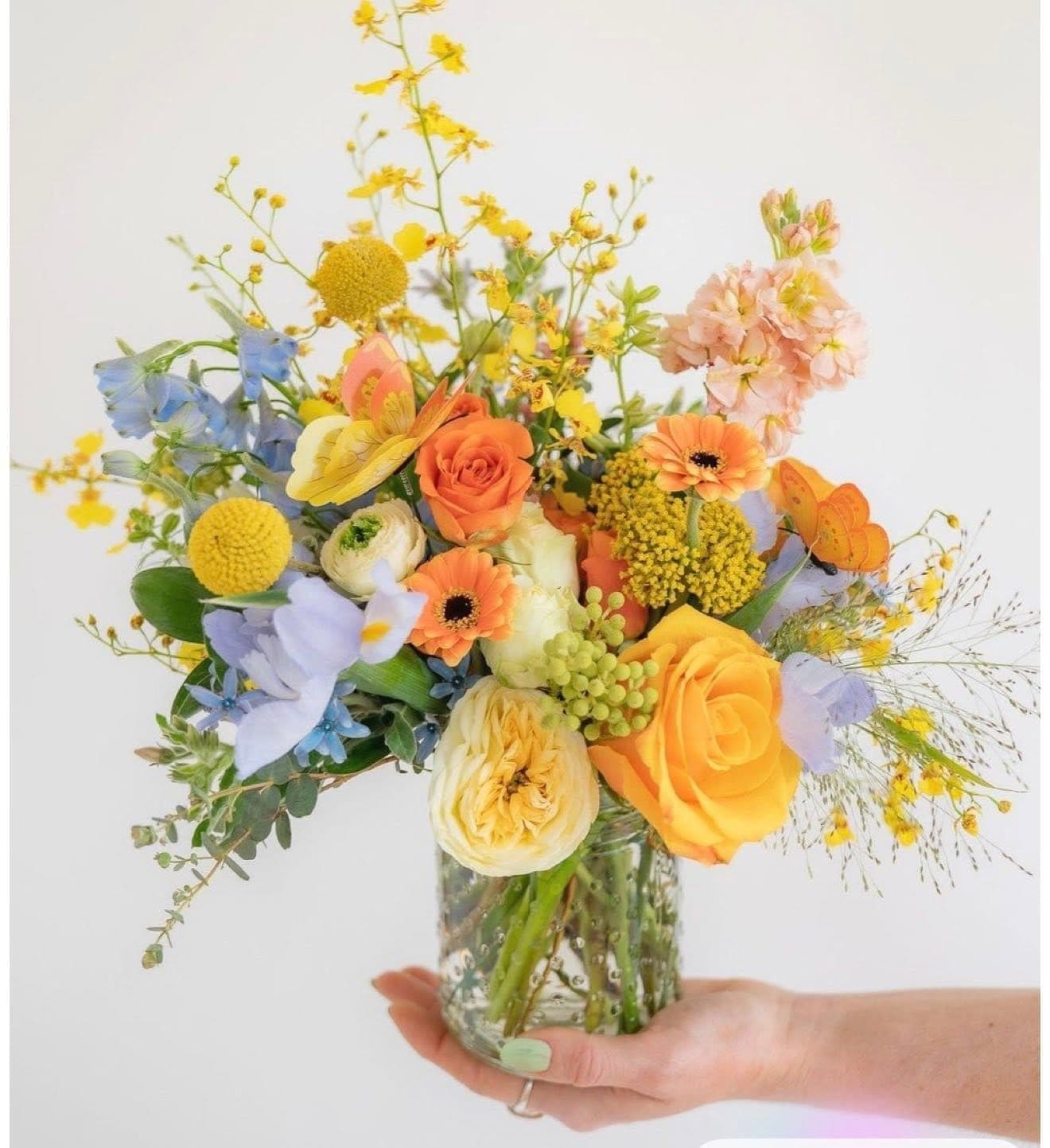 Bright arrangement of orange roses, yellow craspedia, and blue delphinium in a glass vase