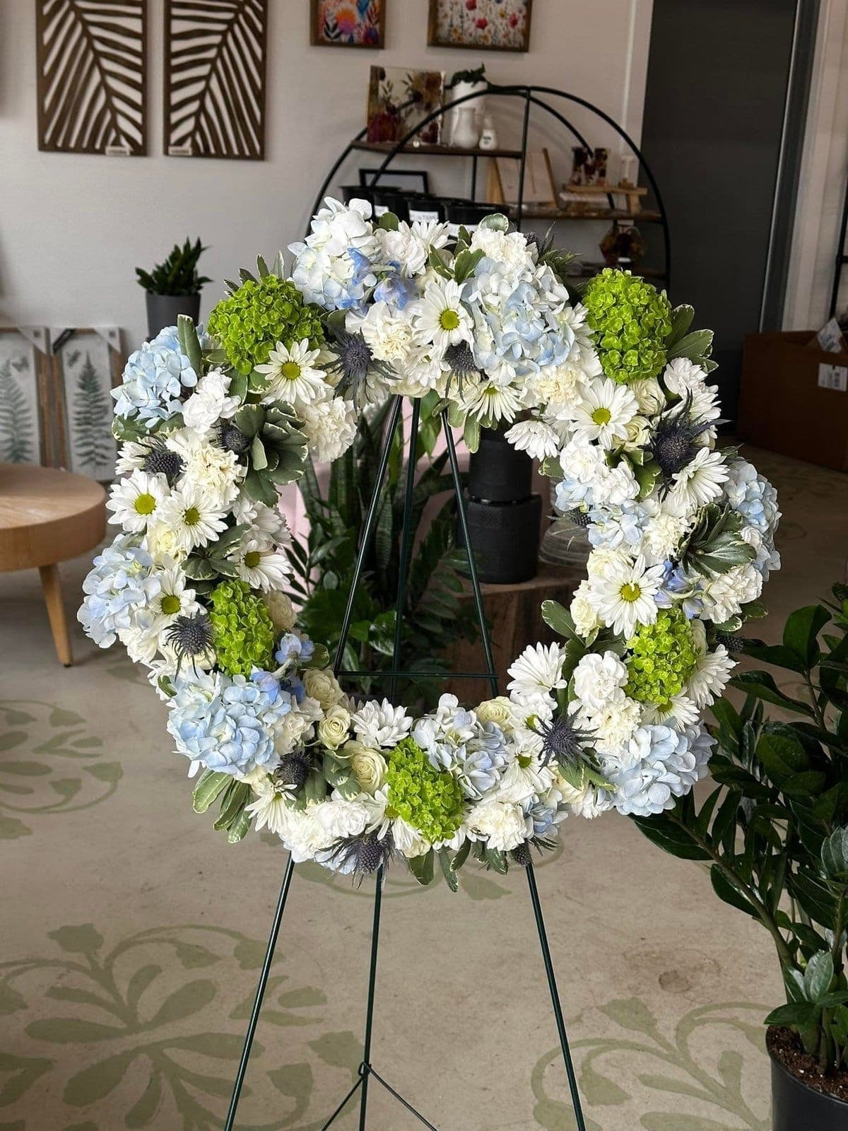 Funeral sympathy wreath of white daisies, blue hydrangea, and green blooms on a standing easel